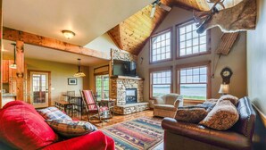 A cozy living room with a stone fireplace, wooden beams, a leather sofa, and a large window overlooking a scenic view.