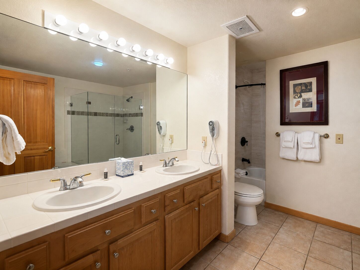 A bathroom with a double sink vanity, a large mirror with lights, a hairdryer, a framed photo, and a shower area.