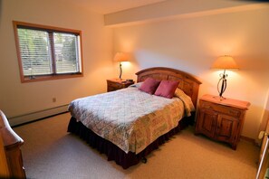 A bedroom with a bed, two bedside tables, a nightstand, a window with blinds, and a lamp on each bedside table.