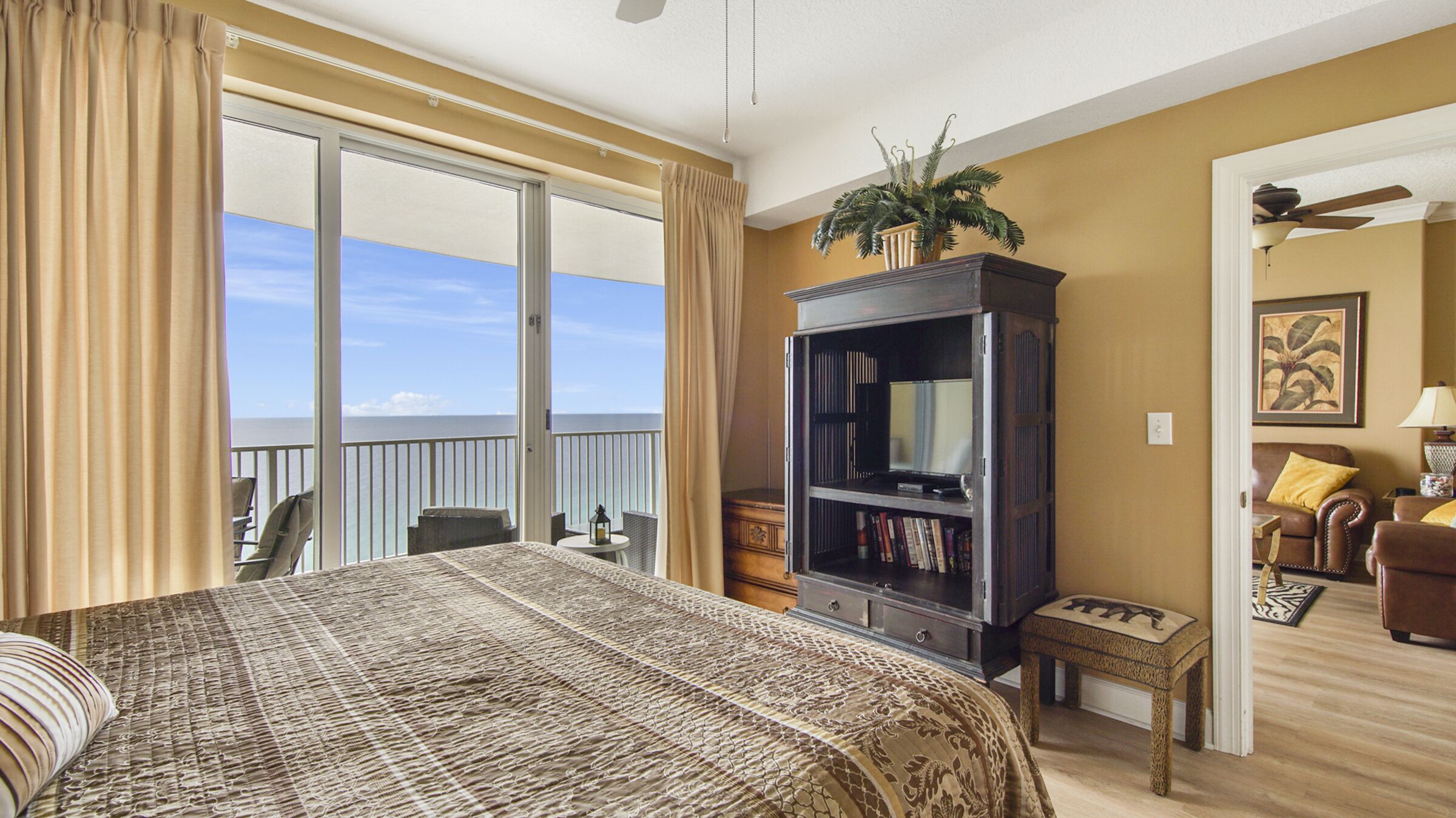 Habitación de hotel con una cama grande, televisión, estantería y balcón con vista al océano.