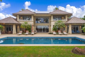 Expansive backyard pool with cushioned lounge chairs, umbrella-shaded table, and tropical palms framing the house facade.