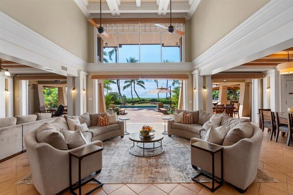 A spacious living area with a high ceiling, large windows, and a view of a pool and palm trees.