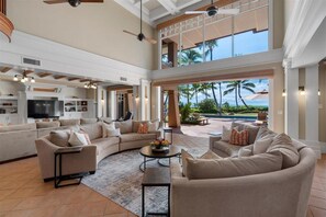 A spacious living room with a large sectional sofa, a coffee table, and a TV in the background. There is a balcony with a view of palm trees and a pool.