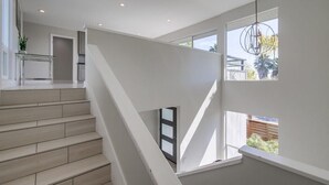Un escalier moderne au design minimaliste, menant à une pièce lumineuse dotée de grandes fenêtres et d’un luminaire suspendu.
