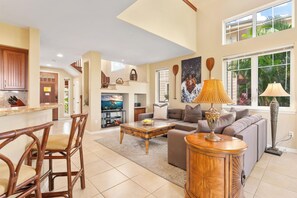 A spacious living room with a large sofa, a coffee table, and a dining area with chairs. There is a kitchen visible in the background.