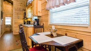 Interior de una cabaña de madera con una mesa de comedor, sillas, una cocina con cafetera y una ventana con persianas.