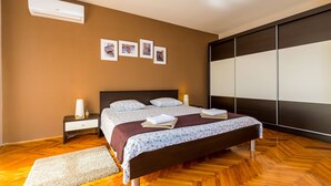Une chambre avec un plancher en bois, un lit recouvert d’une couvre-lit à motifs floraux, une table de chevet avec une lampe et un climatiseur fixé au mur.