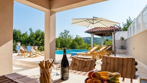 Un espace aménagé au bord de la piscine, avec des chaises longues, une table sur laquelle se trouvent une bouteille de vin et des fruits, et une vue sur la piscine et la végétation environnante.
