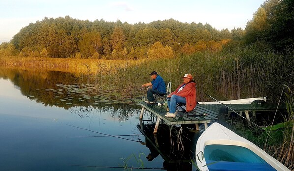 Dos personas pescan en un muelle junto a un lago tranquilo bordeado de juncos y árboles.