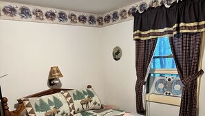 A bedroom with a wooden bed, a nightstand, a lamp, a window with curtains, and a wall with a nature-themed border.