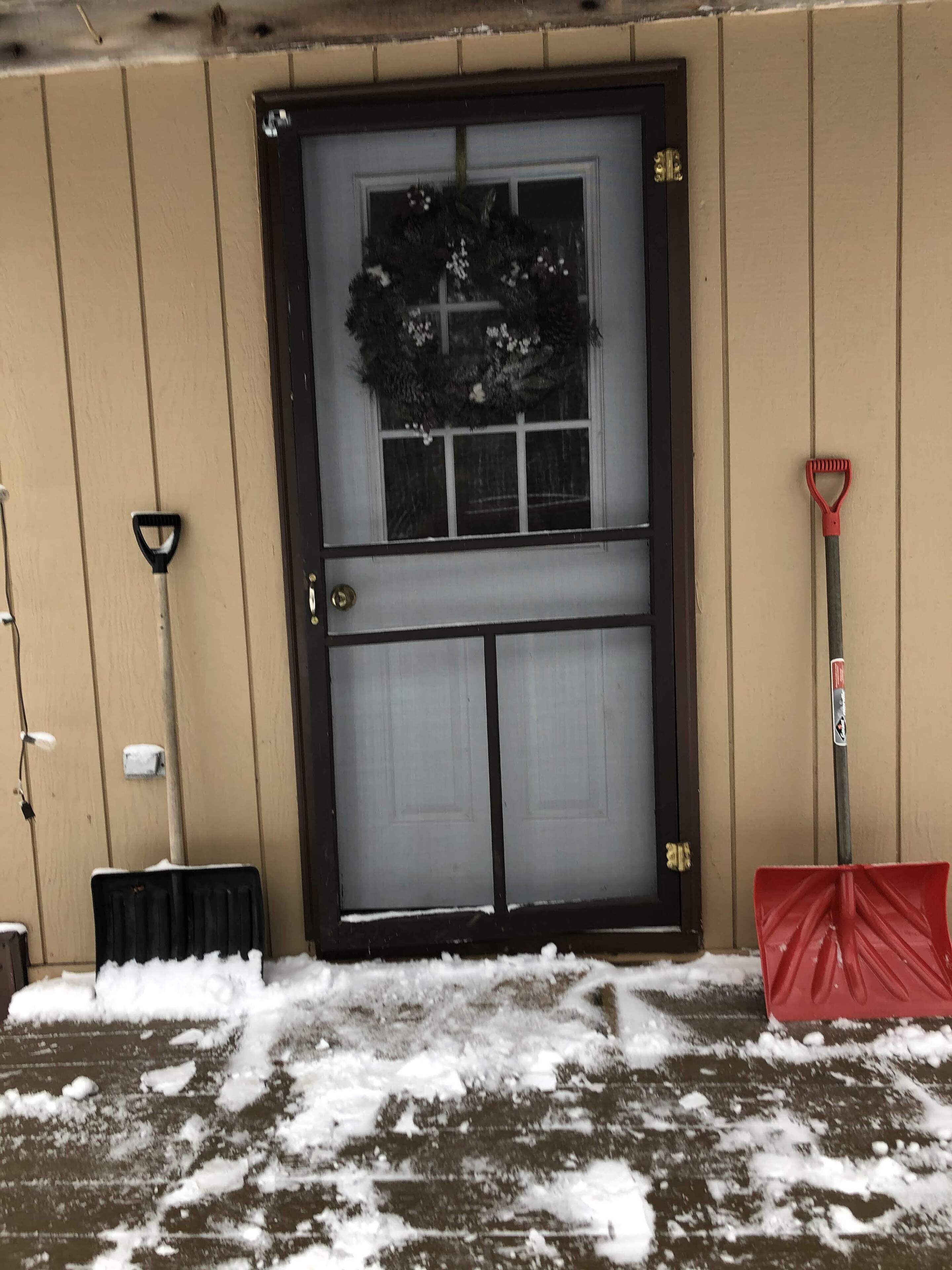 A snow-covered entrance with a door, a broom, and a shovel leaning against the wall.