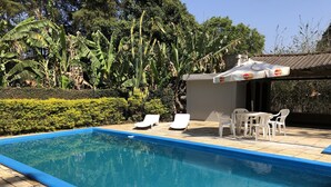 A swimming pool with a tiled deck, surrounded by lush greenery and a shaded area with plastic chairs and an umbrella. - BEAUTIFUL COTTAGE WITH POOL AND LAKE (EXTREMA)