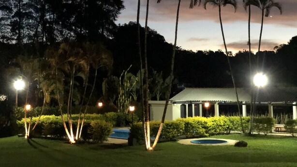 A nighttime view of a well-lit garden with palm trees, a pool, and a building with columns. - BEAUTIFUL COTTAGE WITH POOL AND LAKE (EXTREMA)