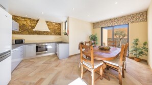 A modern kitchen with a stone wall backsplash, a dining area with wooden chairs, and a view of the outdoors through a large window. - Marim Villas - Villa Flamingo  (Olhão)