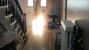 A hallway with a wooden staircase, a door with a stained glass window, and a shoe rack with various slippers.