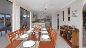 Un comedor con una larga mesa de madera dispuesta para seis personas, rodeada de sillas de madera, con vista a la cocina de fondo.