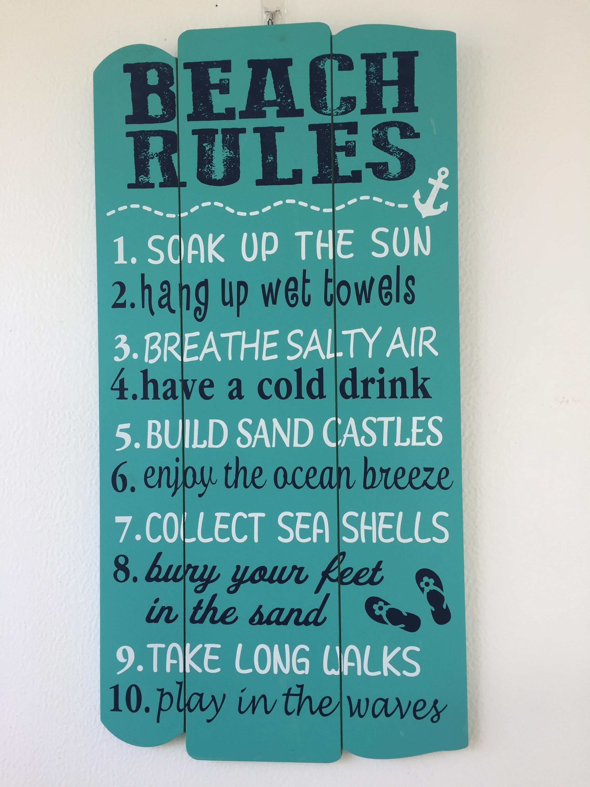 Un letrero azul turquesa con el texto 'BEACH RULES' y una lista de actividades como tomar el sol y construir castillos de arena.