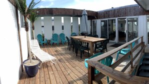 Een houten terras met een tafel en stoelen, een wit hekwerk en een glazen schuifdeur die leidt naar de binnenruimte.