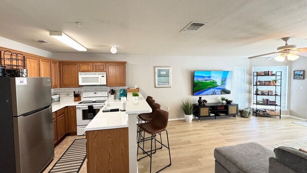Una cocina moderna con armarios de madera, un refrigerador de acero inoxidable y una encimera blanca. Hay un comedor con un taburete de bar y un televisor de pantalla plana. La sala de estar tiene un sofá gris y una estantería.