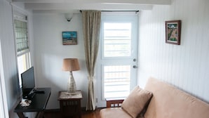Een kamer met een houten bureau, een computer, een lamp, een bank, een deur met luiken, en twee ingelijste foto's aan de muur.