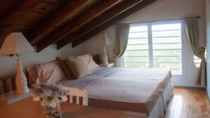 Een slaapkamer met een schuine kap, een groot raam met gordijnen, een bed met kussens, en een nachtkastje met een lamp.