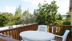 Een houten terras met een witte plastic tafel en stoelen, omgeven door weelderige begroeiing en uitzicht op een waterpartij.