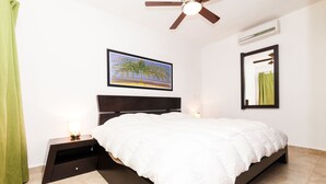 Een slaapkamer met een bed, nachtkastje, plafondventilator, wandairconditioner, een lijst met een afbeelding en een spiegel.