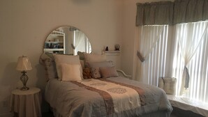 Un dormitorio con cama, mesita de noche con lámpara, espejo sobre la cama y ventana con cortinas traslúcidas.