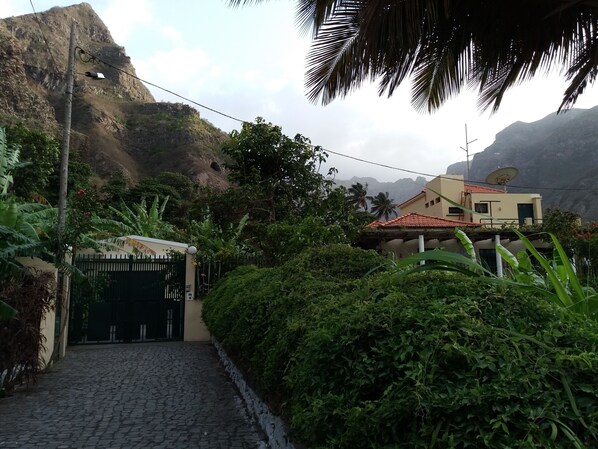 A cobblestone pathway leading to a gated entrance, surrounded by lush greenery and a mountainous backdrop.