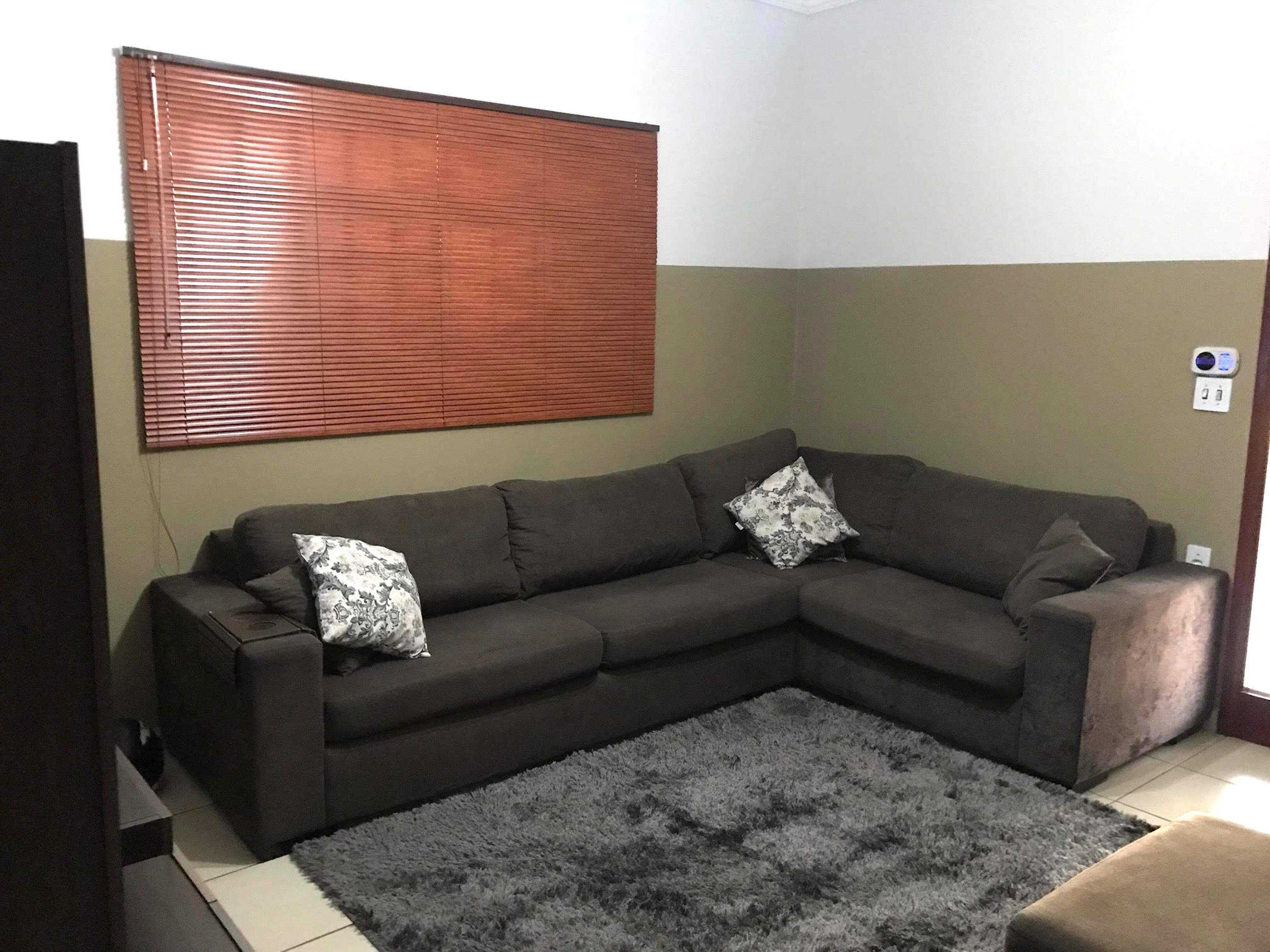 Una sala de estar con un sofá seccional gris oscuro, dos cojines estampados, una alfombra de pelo alto gris y una ventana con persianas.