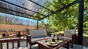 Une terrasse extérieure avec des meubles en bois, une table garnie de nourriture et de boissons, et un espace abrité offrant une vue sur les arbres.