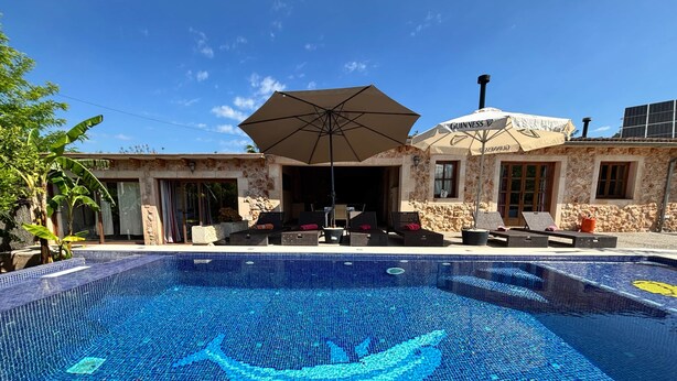 Une piscine ornée d’une mosaïque représentant un dauphin, un espace extérieur aménagé avec des sièges et un bâtiment en pierre surmonté d’un grand parasol.