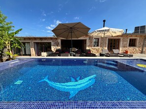 Ein Swimmingpool mit Delfin-Mosaik, Sitzgelegenheiten im Freien und ein Steinhaus mit einem großen Sonnenschirm.