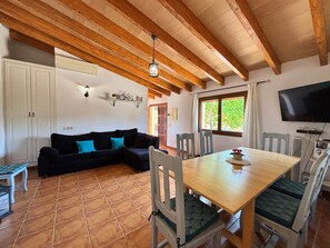 A cozy room with a wooden ceiling, a sofa with teal cushions, a dining table with chairs, a television, and a window with a view of greenery.
