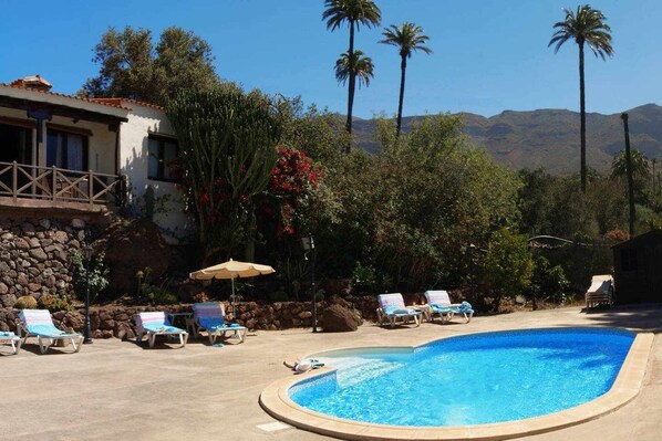 A swimming pool with lounge chairs and umbrellas, surrounded by palm trees and a stone wall. - Casa Rural El Olivar - La Almazara (Valle, El (Santa Lucia De Tirajana))