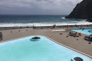 Un espace piscine offrant une vue sur l’océan, une falaise rocheuse et un chemin piétonnier délimité par une clôture.
