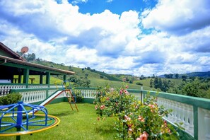 Un parque infantil con toboganes y columpios, rodeado por una cerca verde, con vistas a colinas onduladas y un cielo parcialmente nublado.