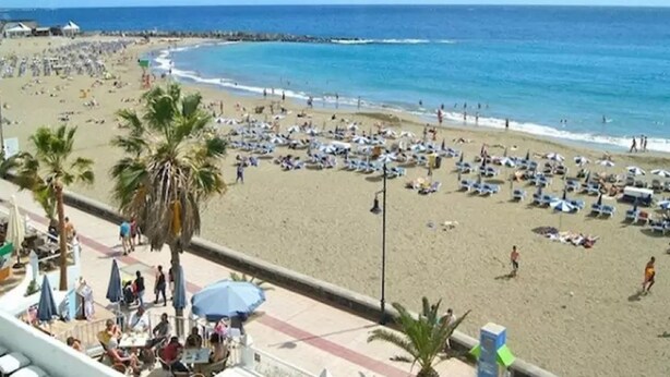 Una playa con numerosas tumbonas y sombrillas, un cielo azul despejado y un mar tranquilo.