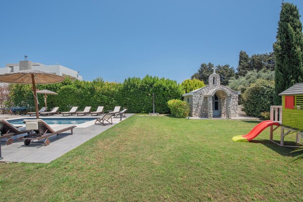 A backyard with a swimming pool, lounge chairs, a small stone building, and a slide.