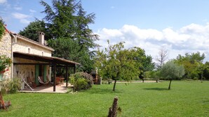 Ein Steinhaus mit überdachter Terrasse, Rasen und Bäumen.