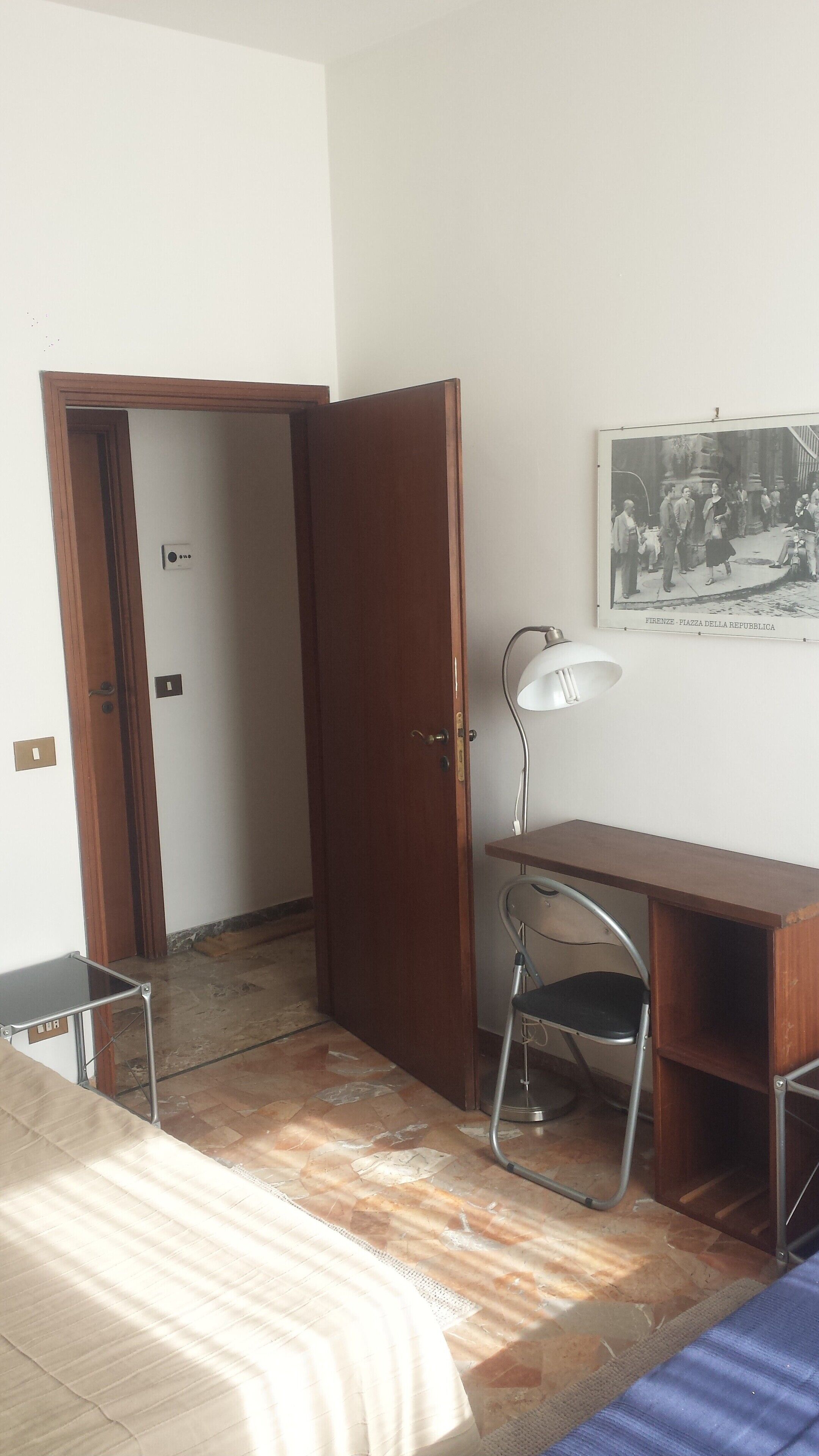 Una stanza con un letto, una scrivania, una sedia, una lampada e un quadro appeso al muro.