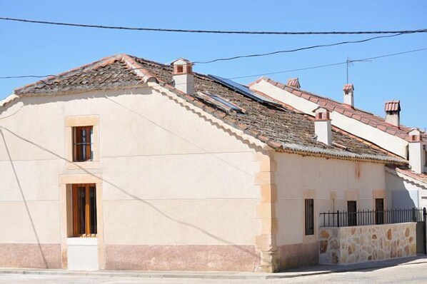 A two-story building with a sloped roof, a small window on the ground floor, and a stone wall on the right side.