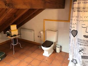 A small, rustic bathroom with a toilet, a radiator, a small table with boxes, and a shower curtain with drawings.