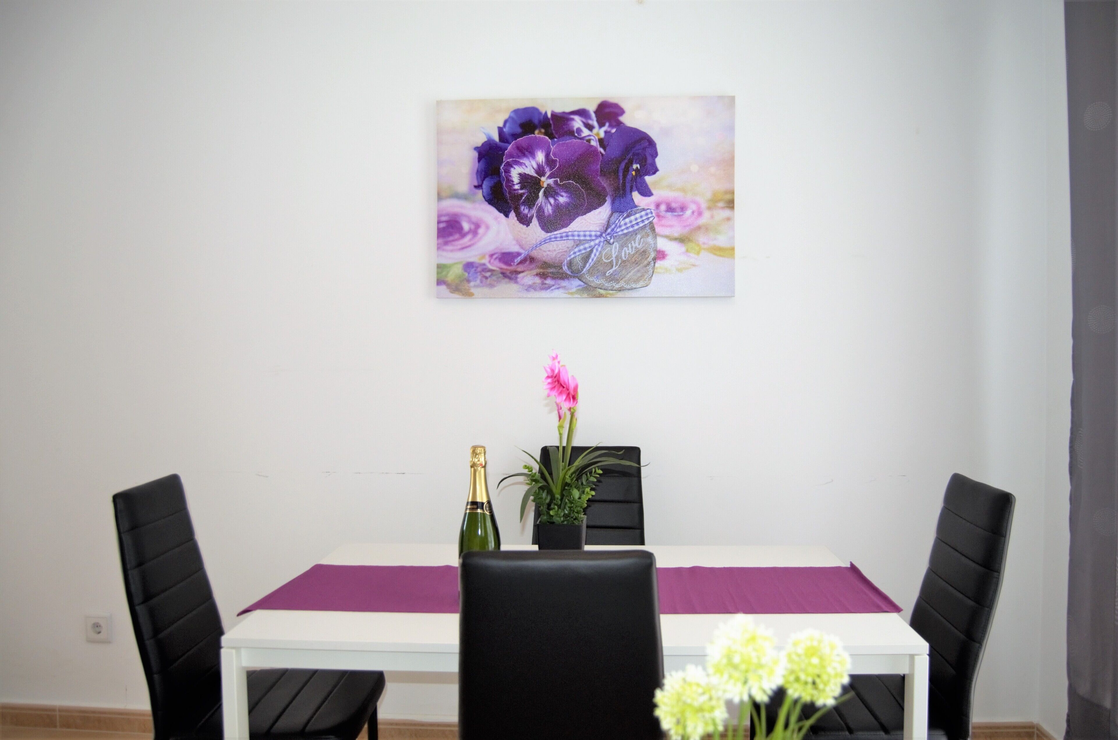 A dining area with a white table, black chairs, a purple table runner, a bottle of champagne, and a vase with flowers.