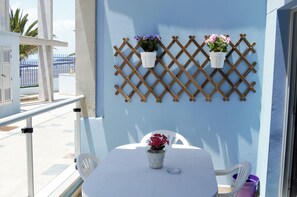 Varanda com mesa branca, duas cadeiras e dois vasos de plantas, tudo em uma parede azul.