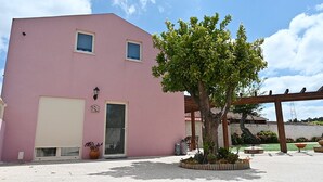 A pink building with two windows, a door, and a small garden area with a tree and potted plants. - Escondidinho Houses - Casa da Eira (Bajouca, Leiria)