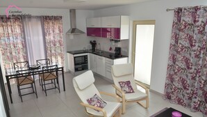A modern kitchen with white cabinets, a black countertop, and a stainless steel range hood. There is a dining area with a glass table and chairs, and a living area with two white chairs and a black coffee table. - Escondidinho Houses - Casa da Eira (Bajouca, Leiria)