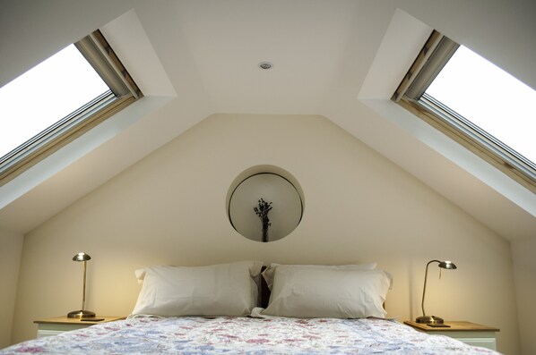Une chambre avec un plafond en pente, des fenêtres de toit, un lit avec une literie à motifs floraux, deux tables de chevet avec des lampes et un miroir fixé au mur.