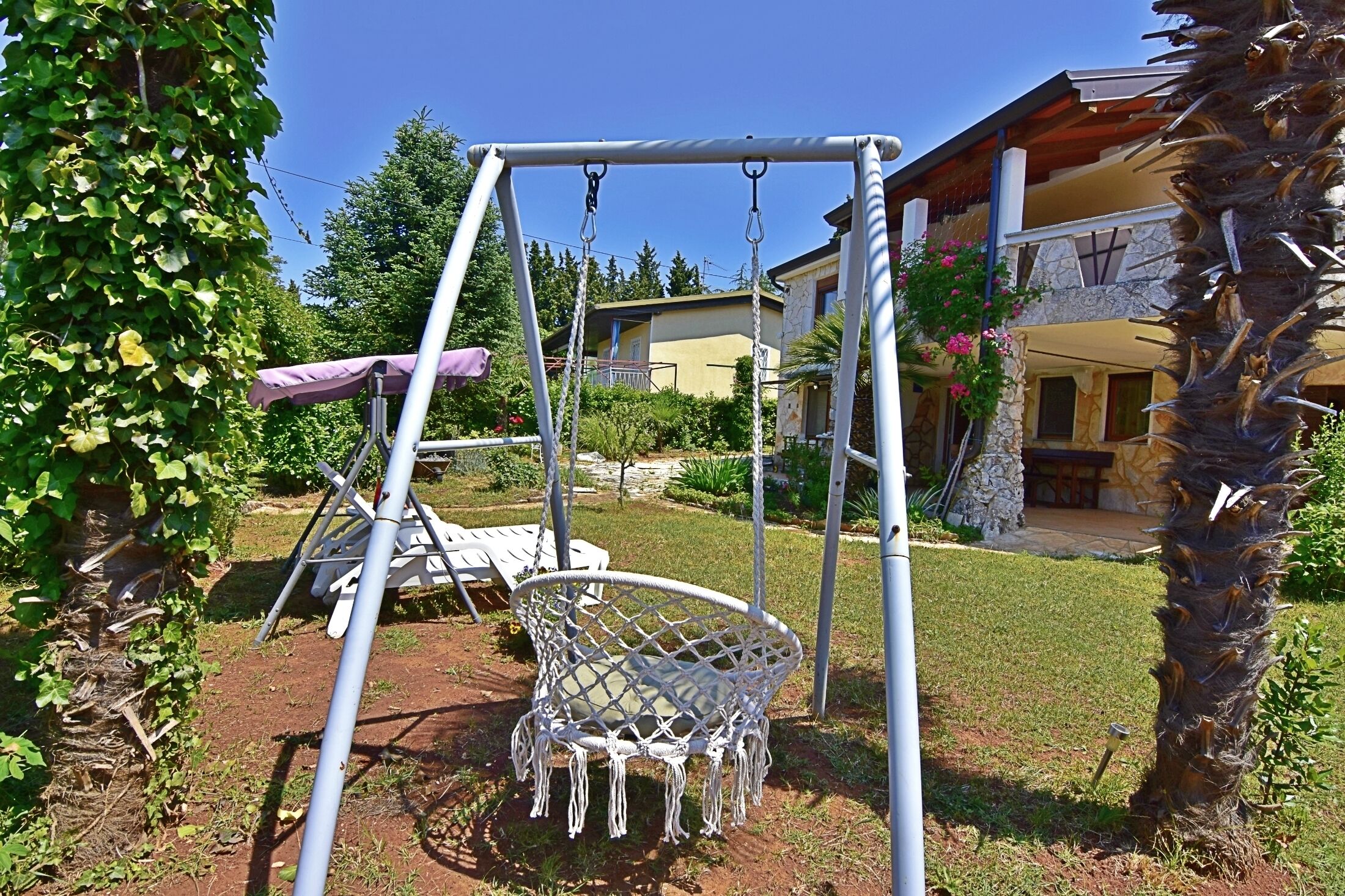 Un columpio con una silla de macramé, un banco cubierto y una casa de piedra al fondo.