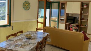 A dining area with a table covered in a patterned tablecloth, wooden chairs, a yellow sofa, and a television on a wooden stand. - House Joseph with beautiful view and private parking (Ravello)